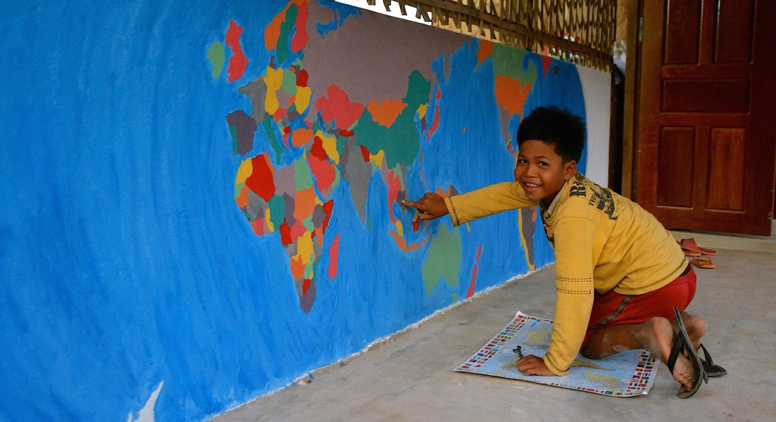 A student at an EDGE Worldwide learning hub in Cambodia pointing to his country on a hand-painted world map mural